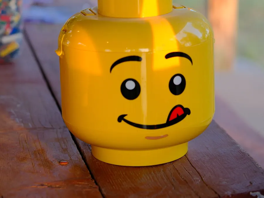 A giant LEGO-head storage bucket with a tongue-out grin, sitting on a wooden picnic table