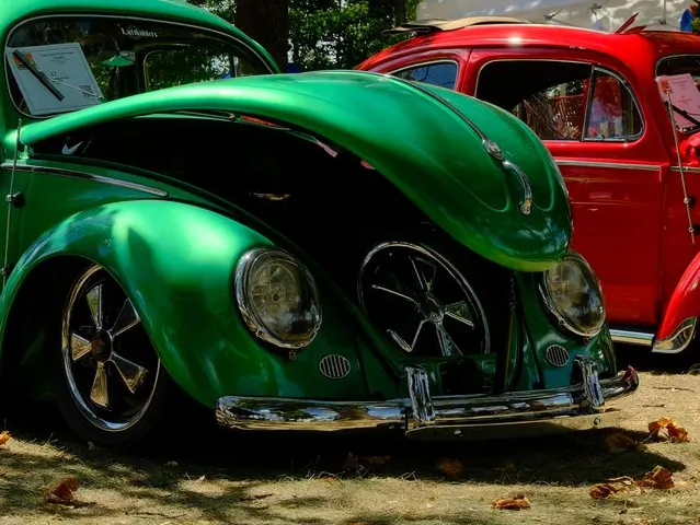 Front end of the green 1955 Legacy Award winner: front trunk lid raised over the spare wheel, round headlights and chrome bumper, with a red Beetle parked beside it