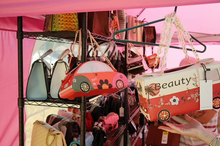Handbags shaped like a Beetle and a VW Bus hanging at a vendor booth under a pink canopy