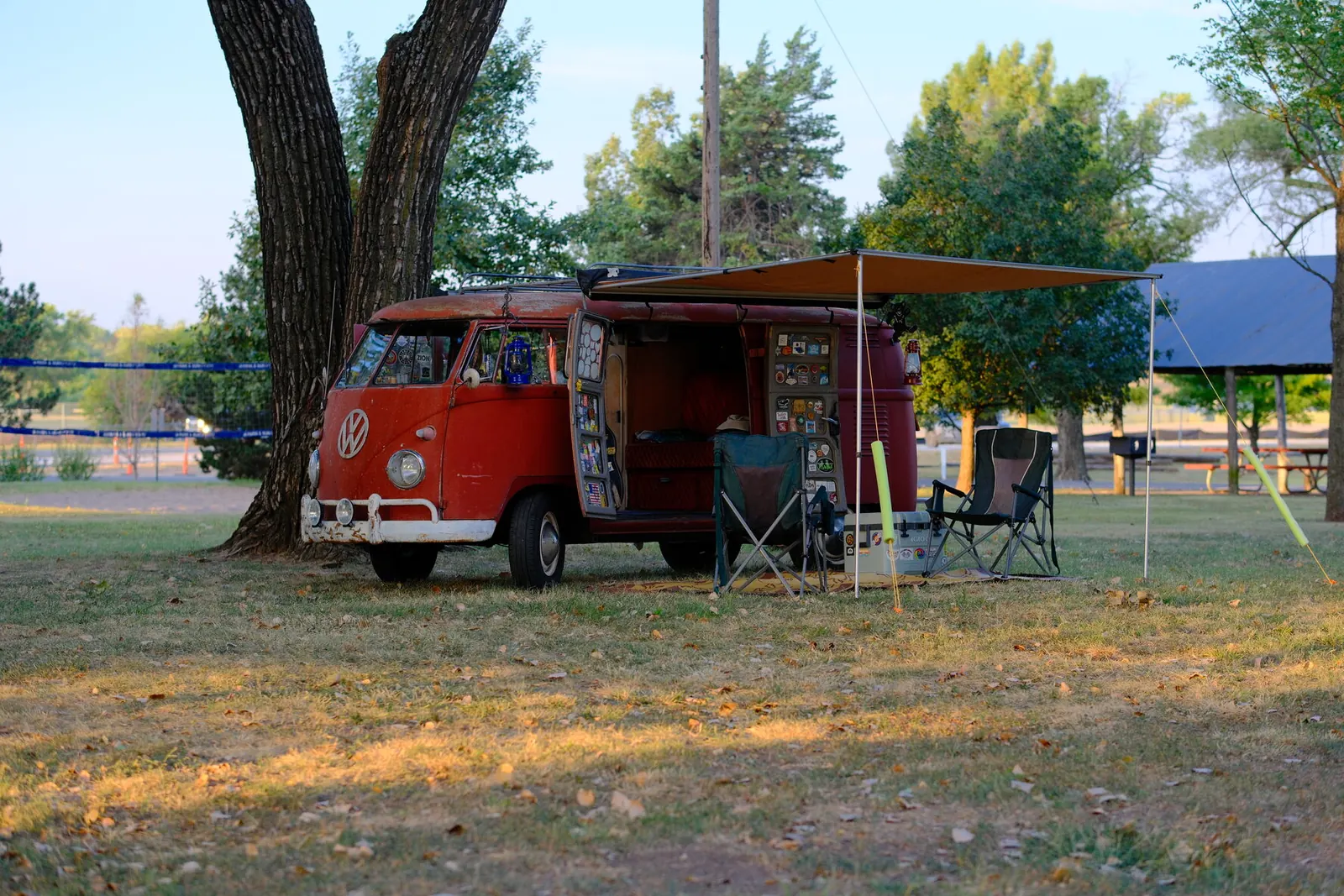 A red split-window VW Bus with its awning out and two camp chairs set up under a tree at Hap McLean Park