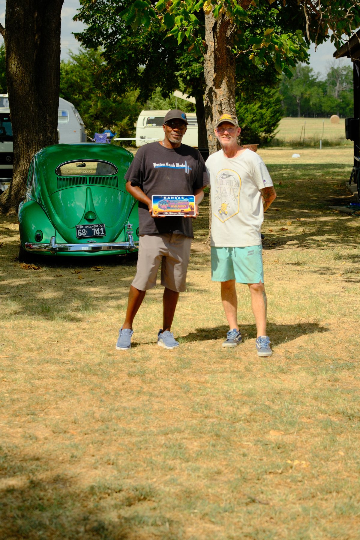 Anthony Bell holding the 2026 Legacy Award plate in front of his metallic green 1955 Beetle at Hap McLean Park